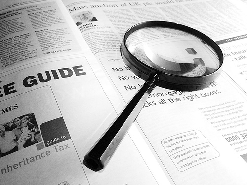 Photograph showing a magnifying glass placed on an open newspaper, focusing on a section about mortgages. The black-handled magnifying glass enlarges text beneath it.
