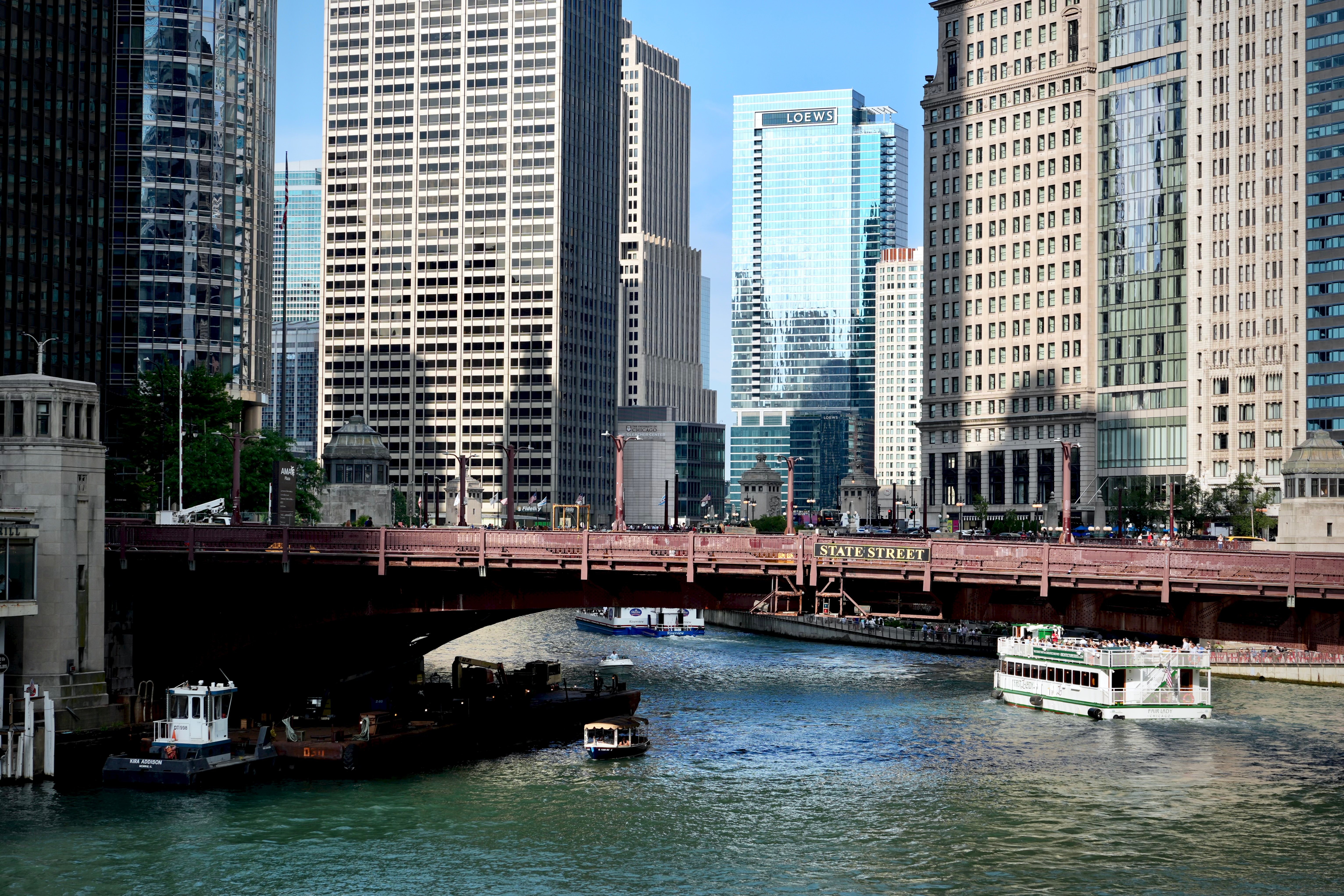 Photograph of Chicago river scene featuring the red steel bridge "State Street" spanning across a waterway with boats and a white tour boat navigating below. Surrounding the river are tall modern and historic skyscrapers, highlighting a bustling cityscape with clear daylight and reflections on glass buildings.